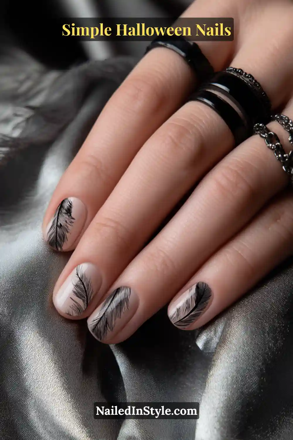 Close-up of squoval nails with a neutral taupe base featuring delicate black feather-like brush strokes along the sidewalls, paired with black chain rings and metallic satin fabric for a dark, elegant Halloween touch.
