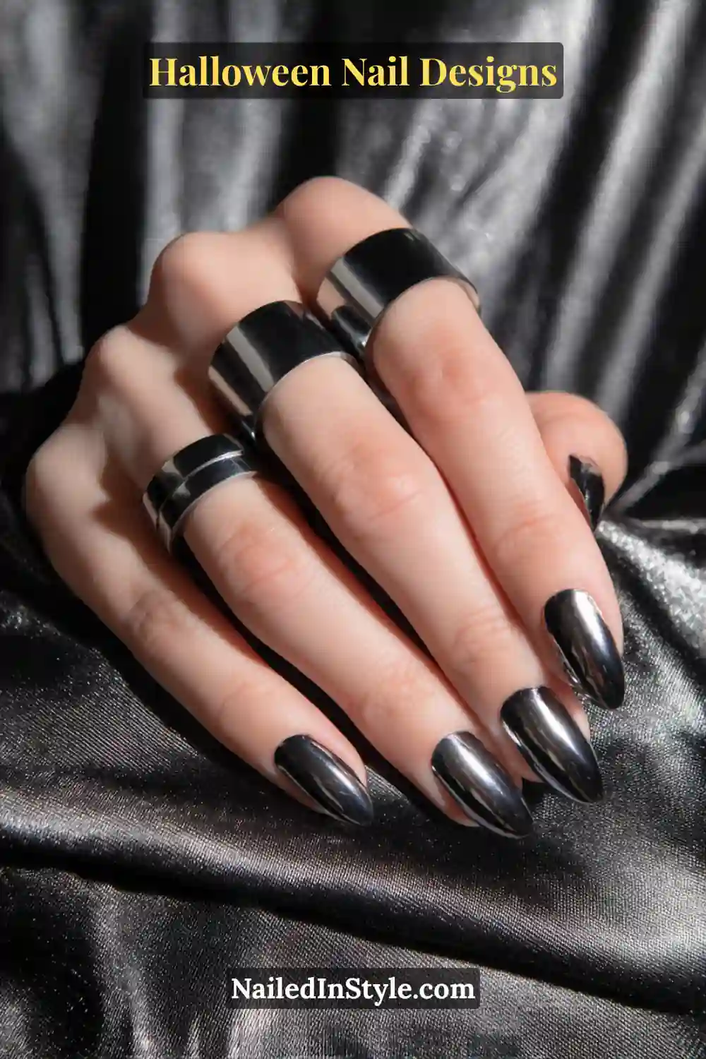Close-up of almond-shaped nails with sleek gunmetal chrome arcs sweeping diagonally across a glossy black base, styled with bold silver rings on a metallic background.