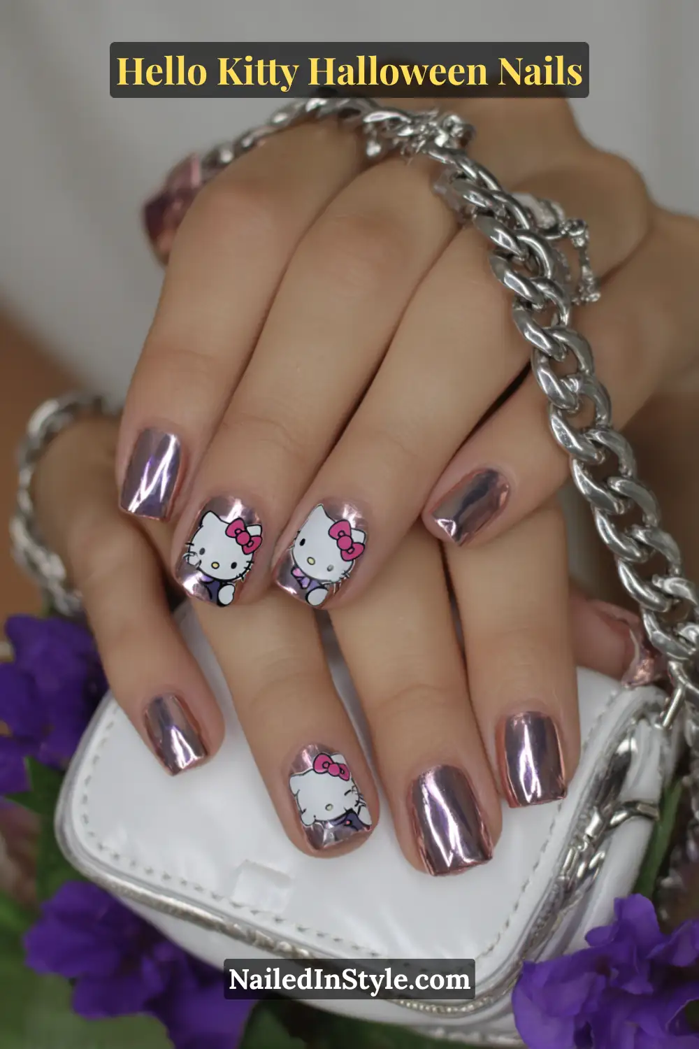Medium square nails with a rose-chrome mirror finish framing a sheer base, featuring playful Hello Kitty art for a chic yet whimsical Halloween look. Styled with a silver chain and rose clutch for a polished, glam touch.