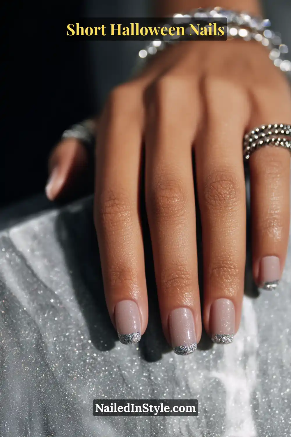 Short square nails with soft nude base and a delicate line of micro silver pinpoints tracing one sidewall, glossy pinpoint sparkle finish; styled with silver beaded rings and shimmering marble surface, elegant Halloween nails for all skin tones.