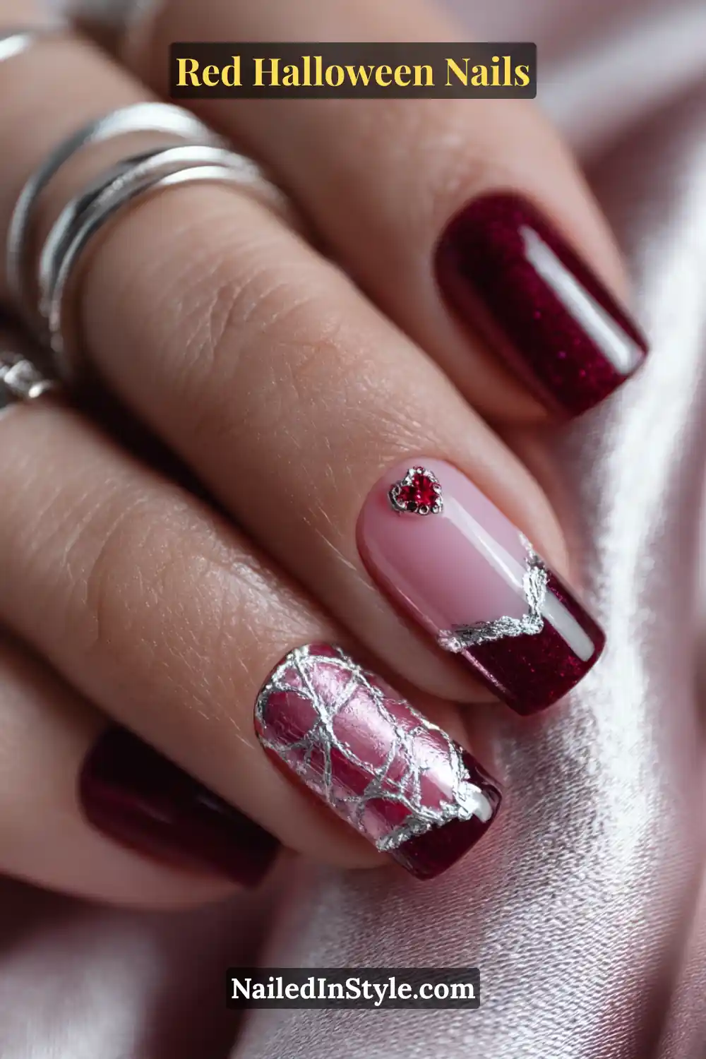 Red Halloween Nails Medium square nails with a mix of rose nude and ruby red tones, featuring micro silver cuffs, a faint raised heart-web design, and a tiny red gem accent; styled with silver stacked rings on a satin background; complements fair to deep skin tones.