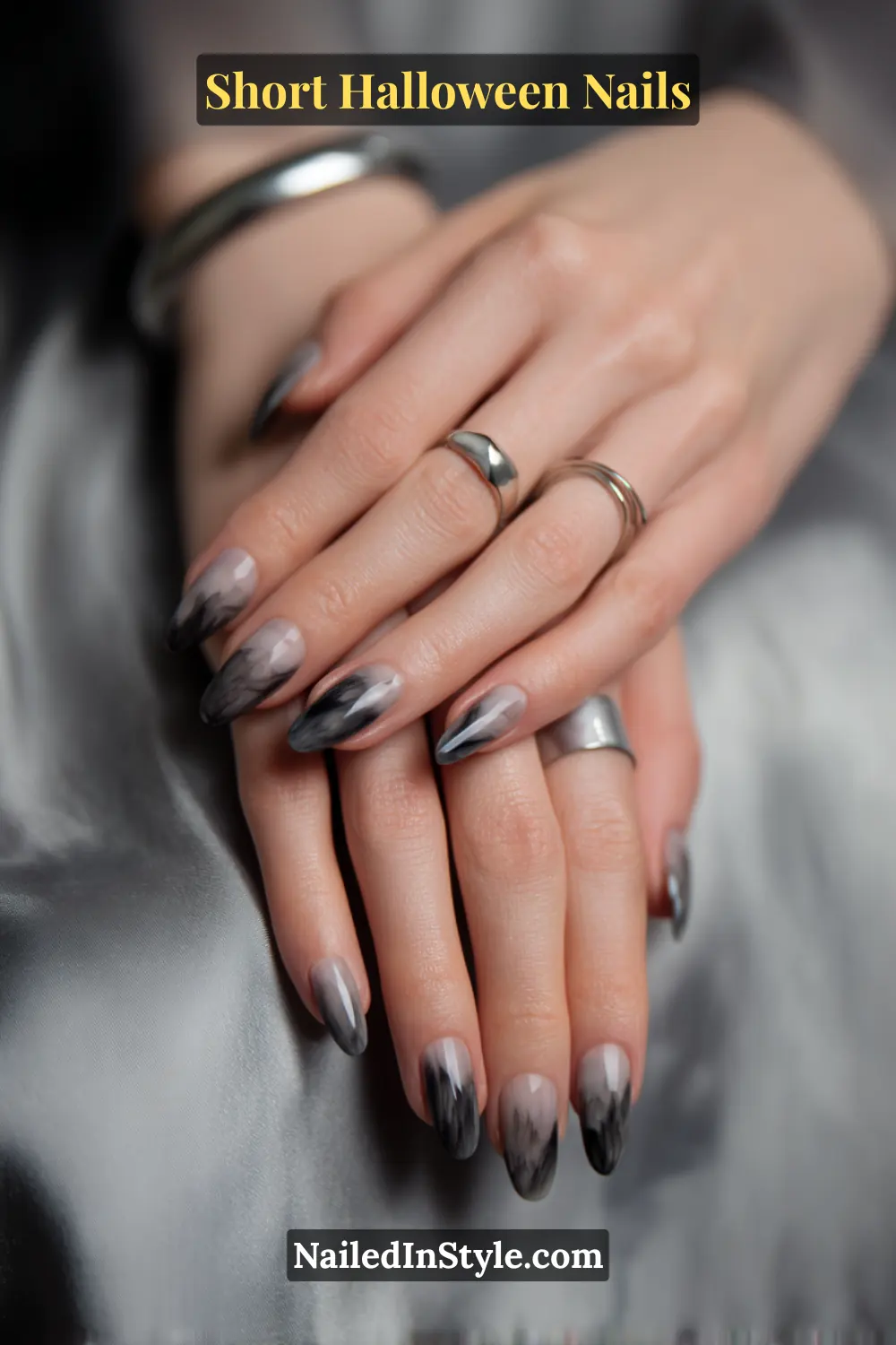 Short oval nails with a sheer nude glaze and ultra-fine charcoal flicks extending from mid-nail to tip, creating a feathered motion effect; styled with sleek silver rings and gray satin fabric, modern Halloween nails for fair and medium skin tones.