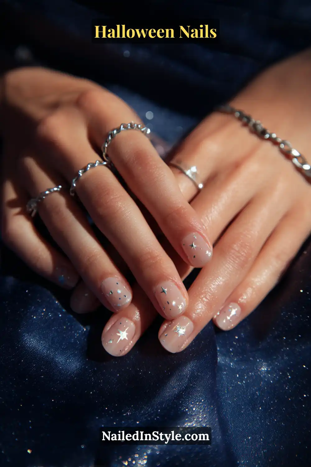 Short square nails in a sheer nude base scattered with tiny sterling star pinpoints made from foil or crystals, glossy pinpoint sparkle finish; styled with silver chain and twisted rings against a deep navy satin fabric, stargazer-chic Halloween nails for all skin tones.