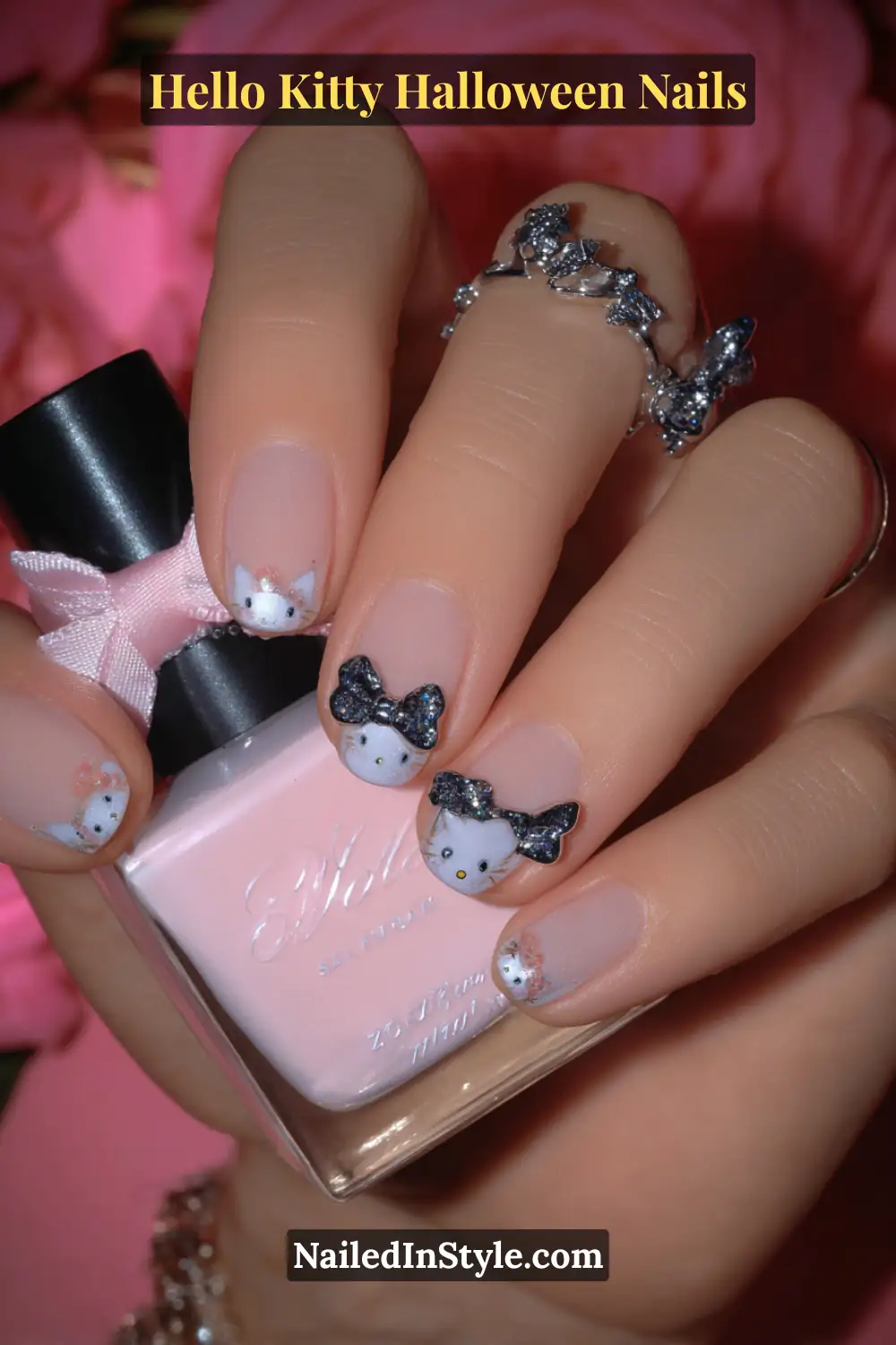 Short squoval nails with a petal pink gloss base, featuring 3D candy-textured Hello Kitty bow charms in silver and black. Styled with dainty rings and a soft pink nail polish bottle for a girly Halloween look.