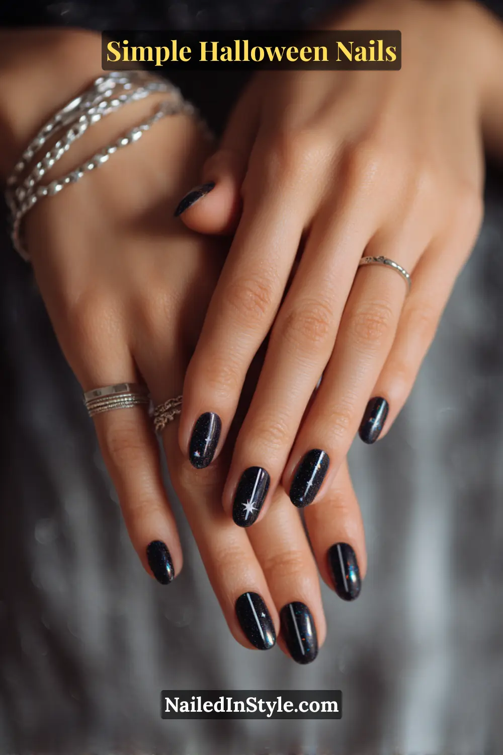 Close-up of short oval nails with glossy black polish featuring a fine vertical graphite line and a tiny silver star at the apex, styled with layered silver rings and bracelets on a dark shimmering fabric.