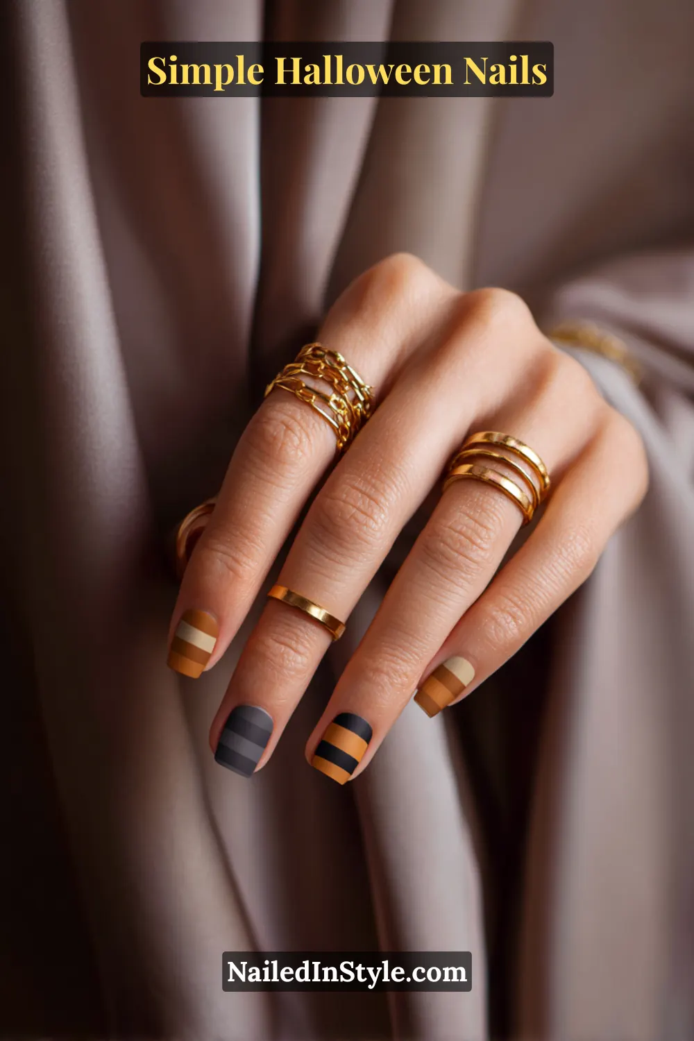 Close-up of short square nails featuring three stacked horizontal stripes in charcoal, amber, and ivory at the tips, styled with layered gold rings and soft taupe drapery for a modern fall look.