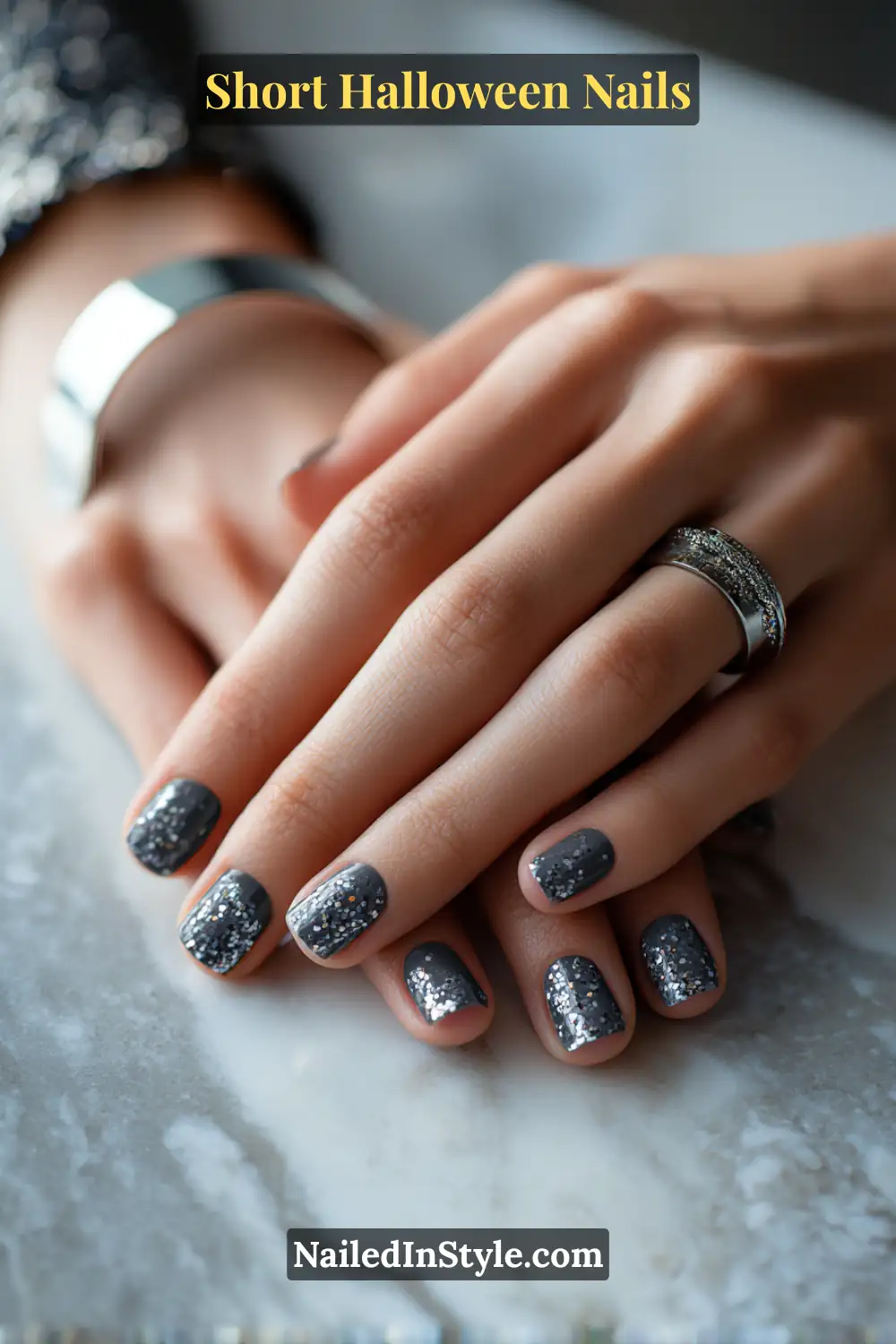 Short square nails in a deep graphite base featuring sparse hematite and sterling confetti accenting the cuticle corners, paired with silver statement rings on a marble surface for a festive yet refined Halloween nail style.