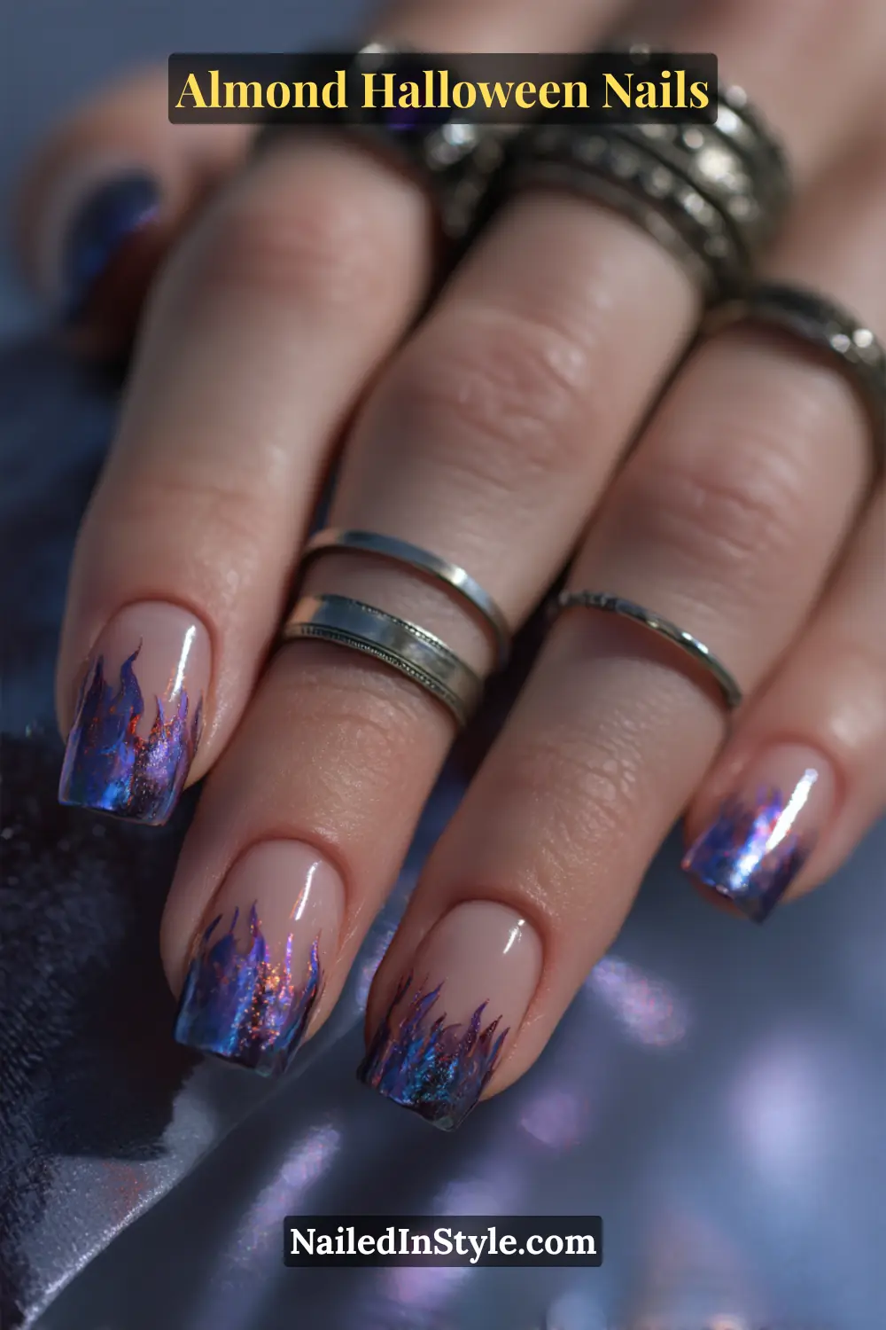 Close-up almond nails with nude base and violet-to-indigo foil fire streaks at glossy tips, shimmering with metallic reflections, Almond Halloween Nails.