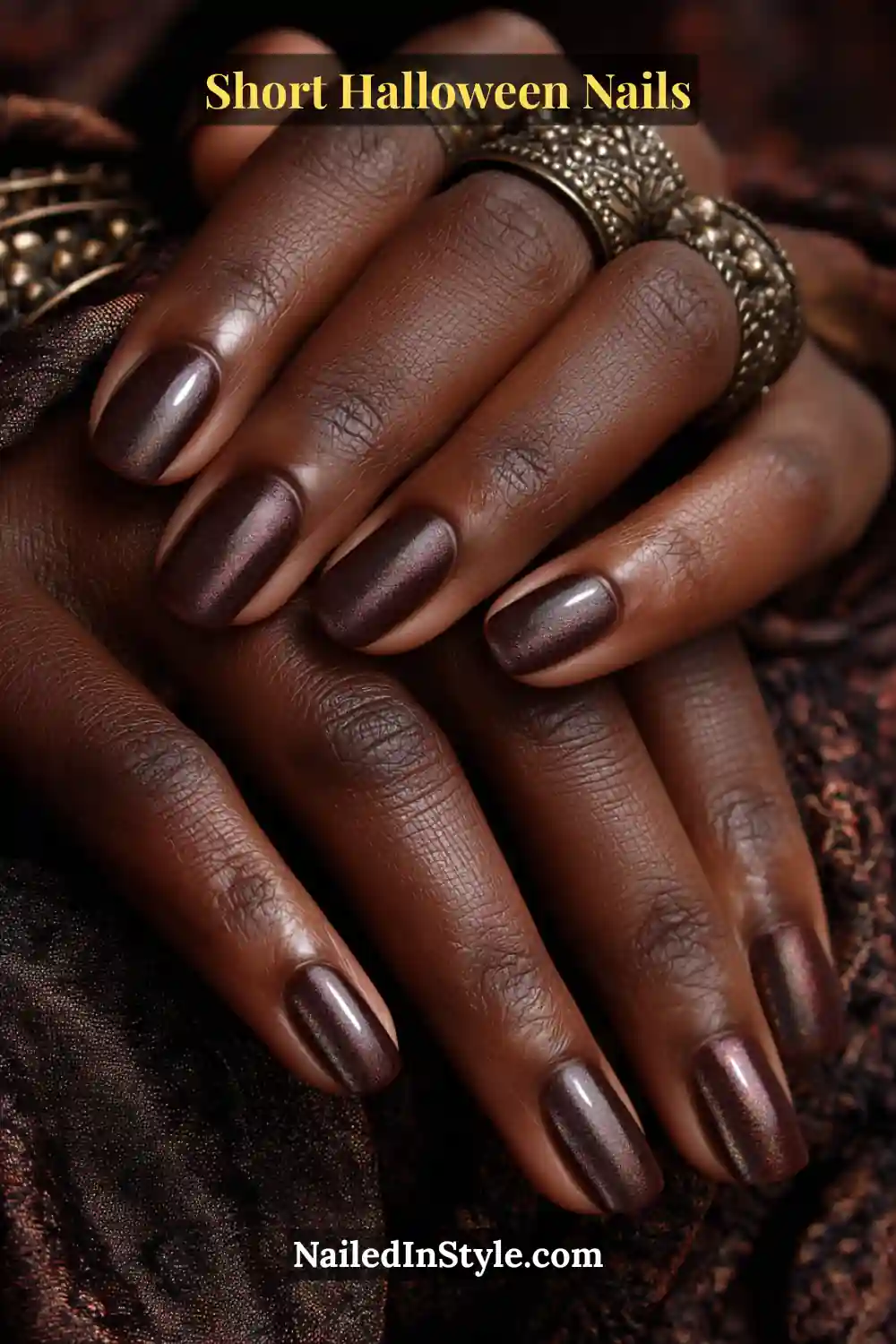 Short squoval nails in rich espresso brown with a subtle iridescent beetle shift centered at the apex; styled with antique bronze rings and dark textured fabric, luxurious short Halloween nails for deep and medium skin tones.