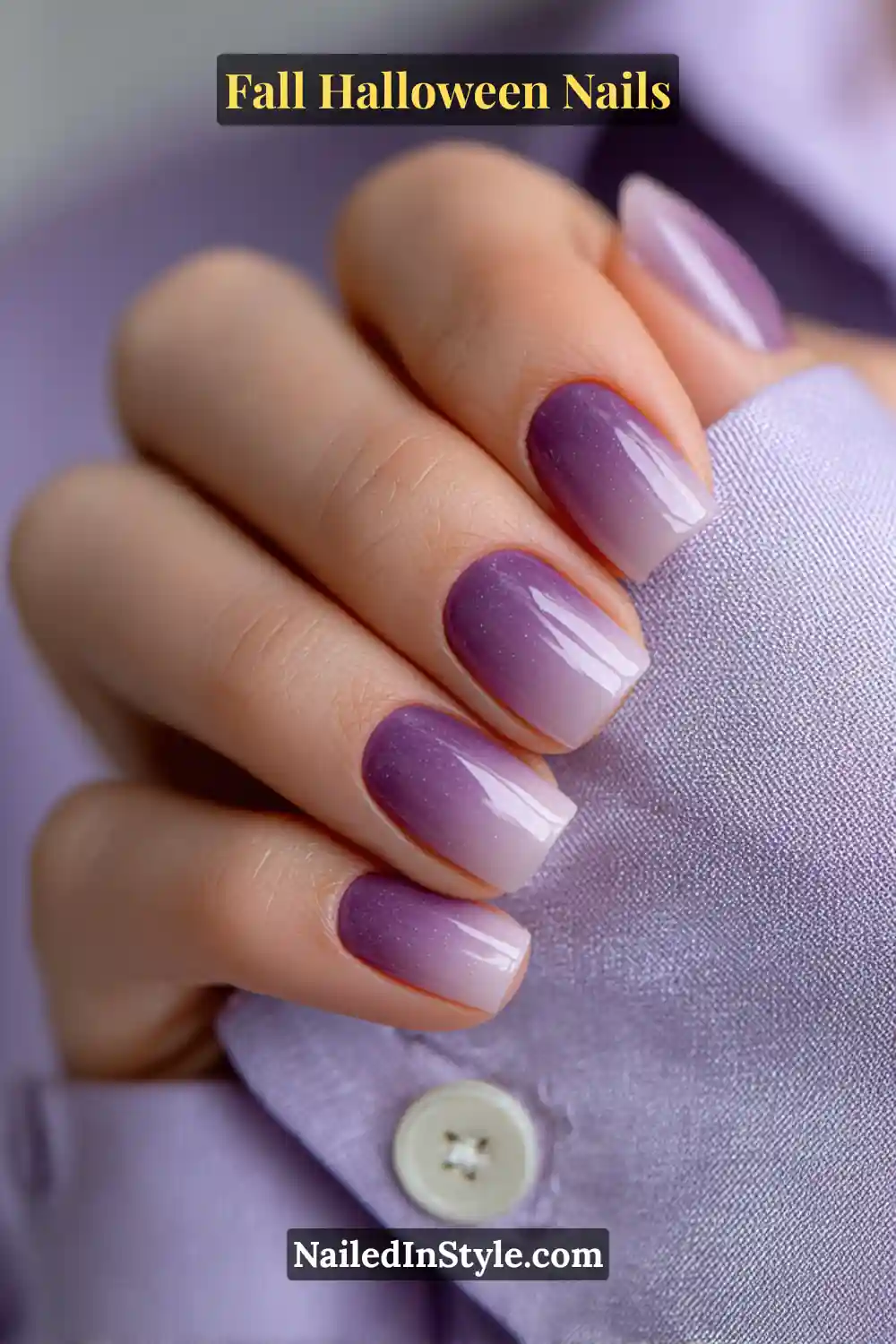 Short squoval nails in a smoky violet-to-ash ombré with a satin-gloss finish, styled against a lilac blouse for a polished autumn-Halloween transition look.