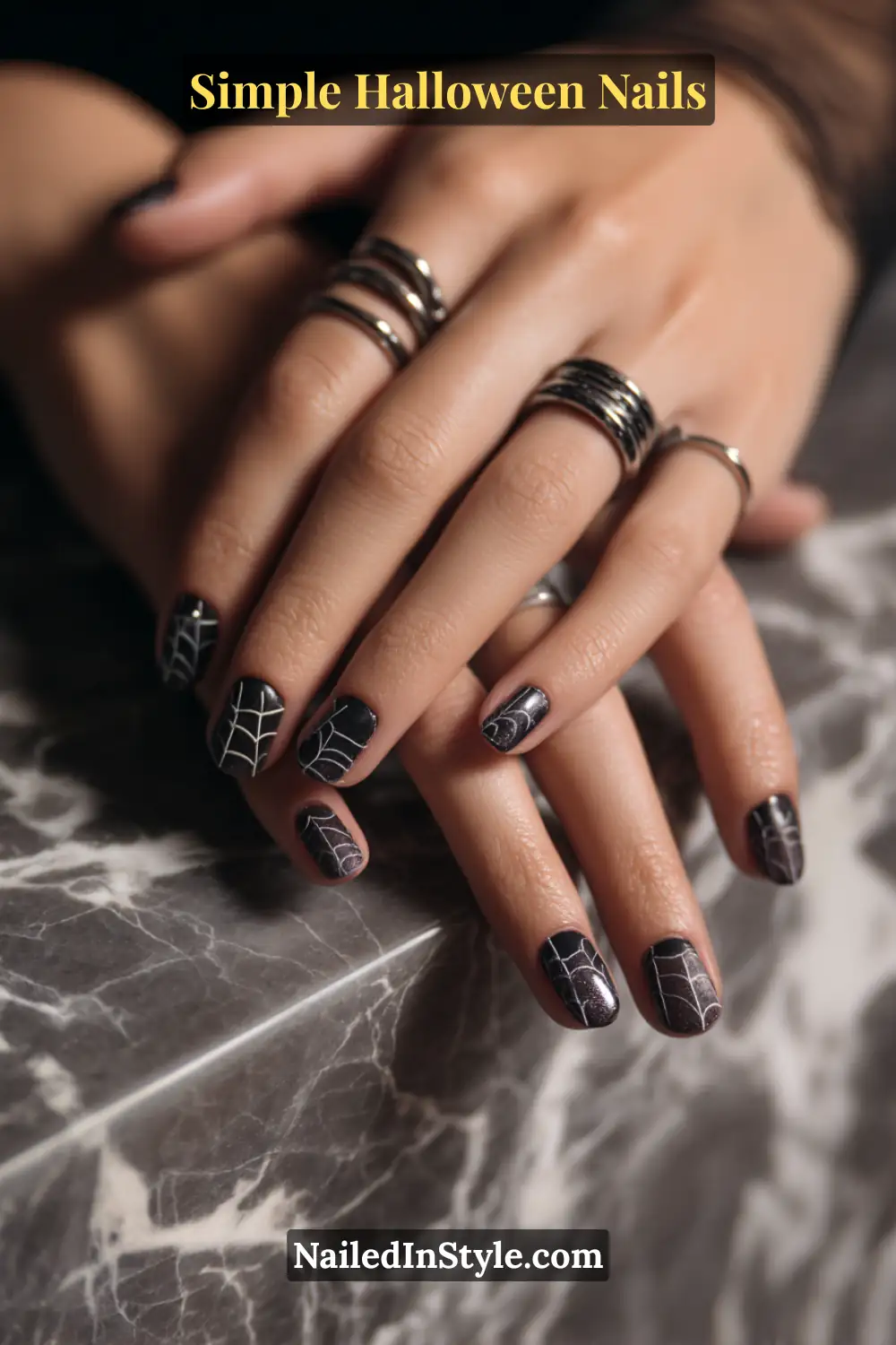 Close-up of short squoval nails painted in glossy black with fine white web patterns tucked into the corners, styled with silver rings and a marble surface for an elegant Halloween aesthetic.
