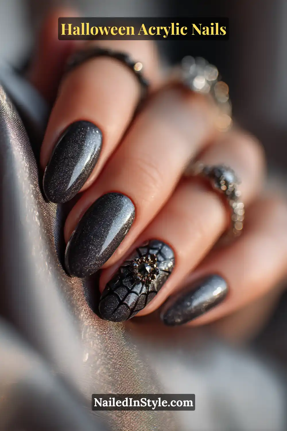Close-up of oval acrylic nails featuring metallic spiderweb designs anchored with onyx gemstone medallions on a shimmery charcoal base, styled with dark silver gothic rings over satin fabric.