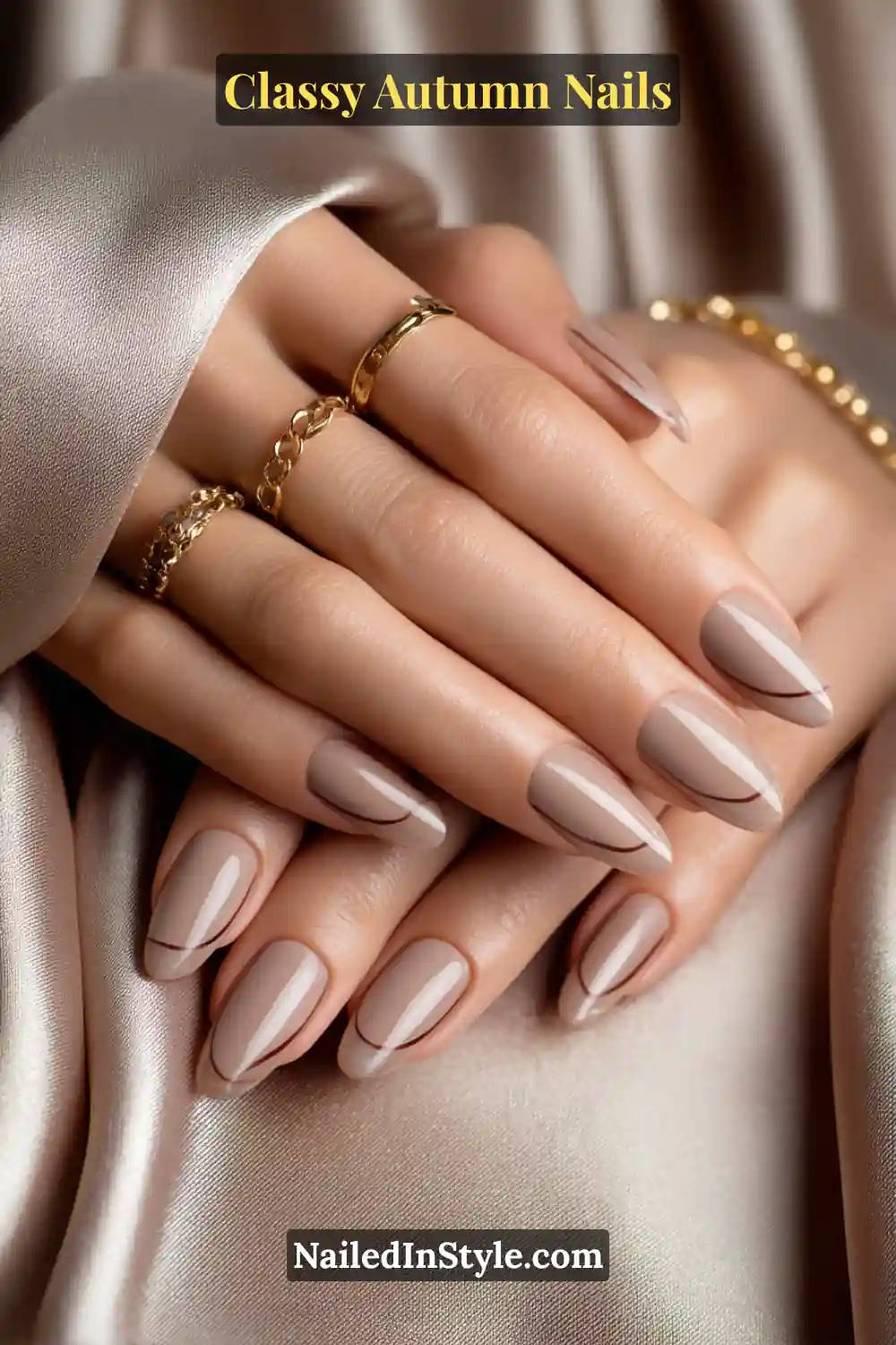 Almond nails in milk-tea beige with thin sable-brown bowlines slightly off-center, paired with gold rings and satin fabric.
