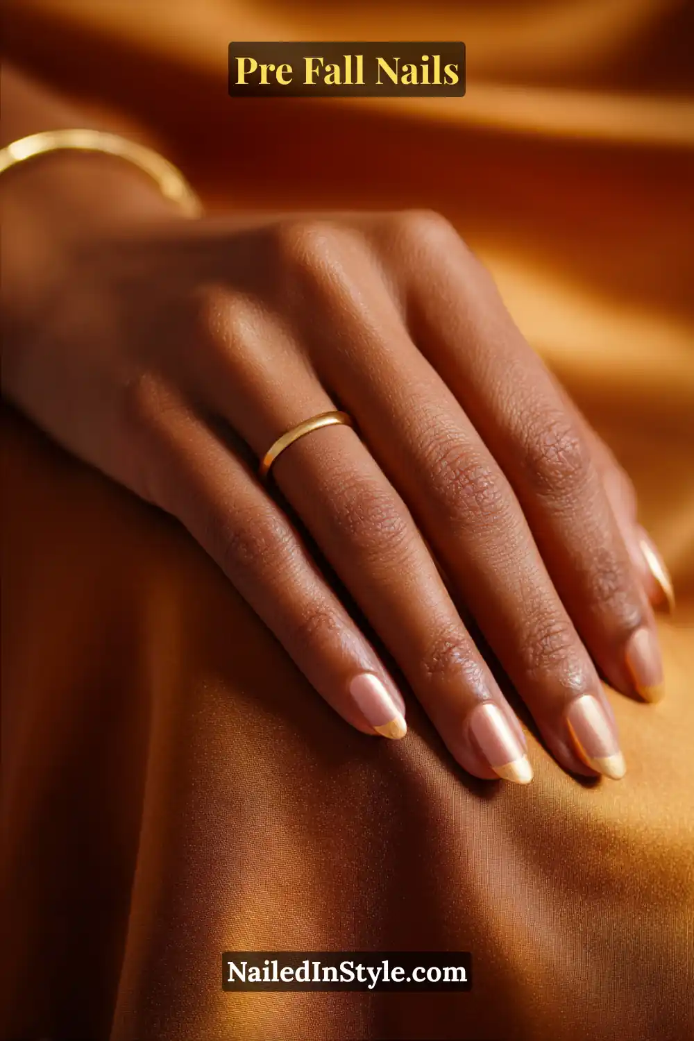 Oval nails with a subtle cider-gold half moon at the cuticle, styled on deep skin with gold jewelry against a bronze satin background.