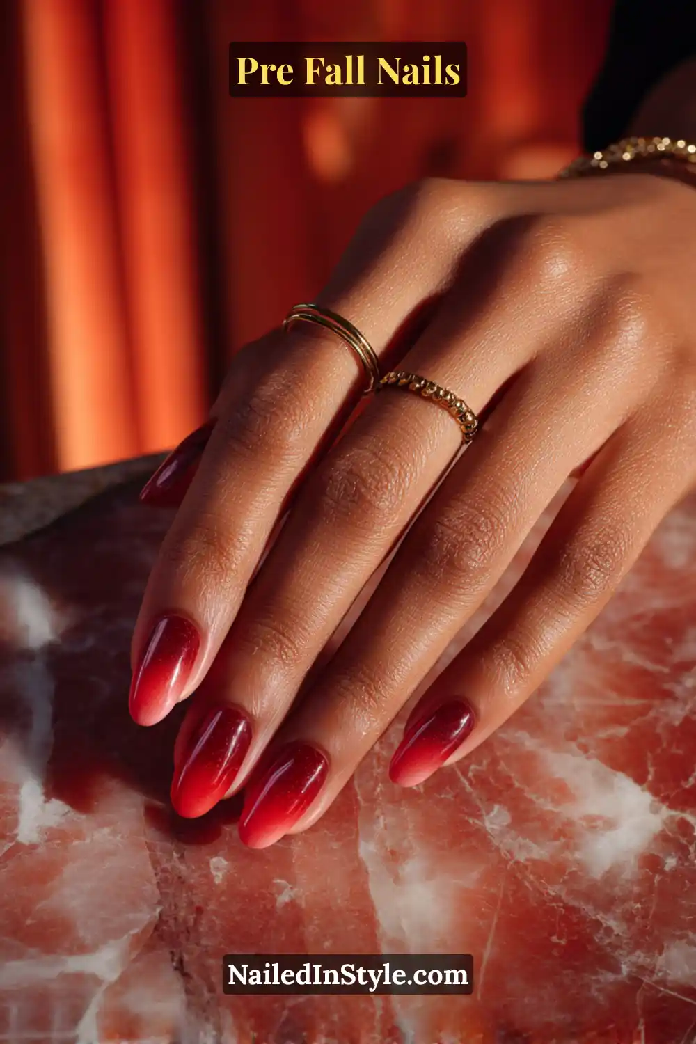 Almond nails in a cranberry and apple-red side-to-side ombré gradient, styled with gold rings on deep skin against a warm marble surface.