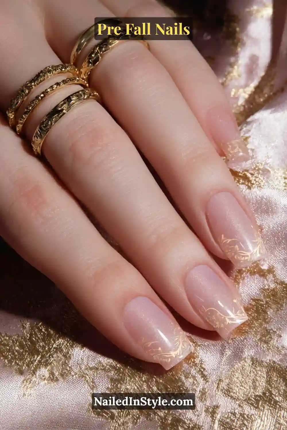 Short square nails in translucent maple nude with delicate gold filigree detailing near the cuticle, styled with ornate gold rings on metallic fabric.