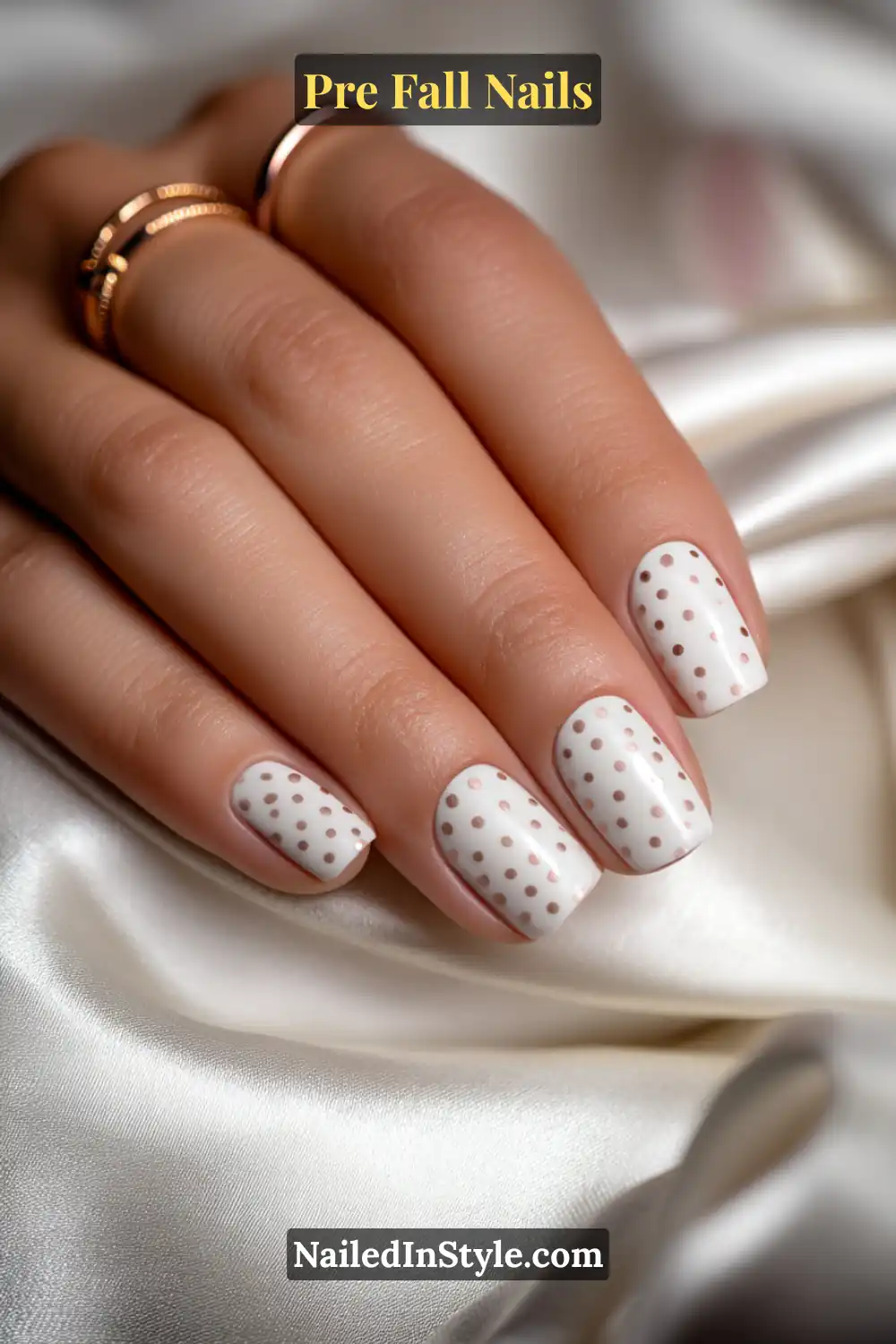 Short square nails with a porcelain white base and evenly spaced mocha micro-dots, styled with gold rings on soft ivory satin fabric.