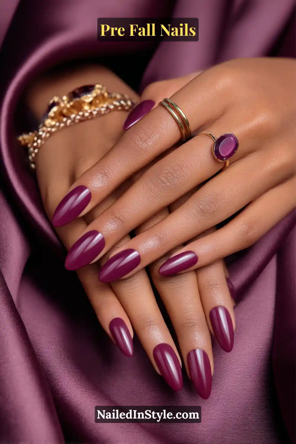 Almond nails painted in rich plum with a sleek crescent accent along one sidewall, styled with gold and plum gemstone rings on deep plum satin fabric.