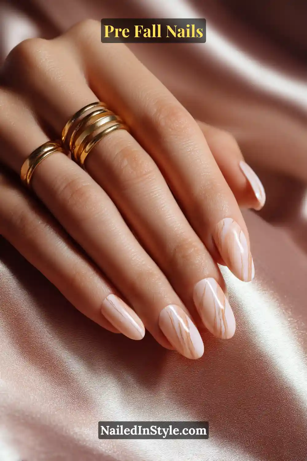 Oval nails with a latte base overlaid by soft sienna-toned wisps resembling fine marble veining, paired with gold rings on a satin backdrop.