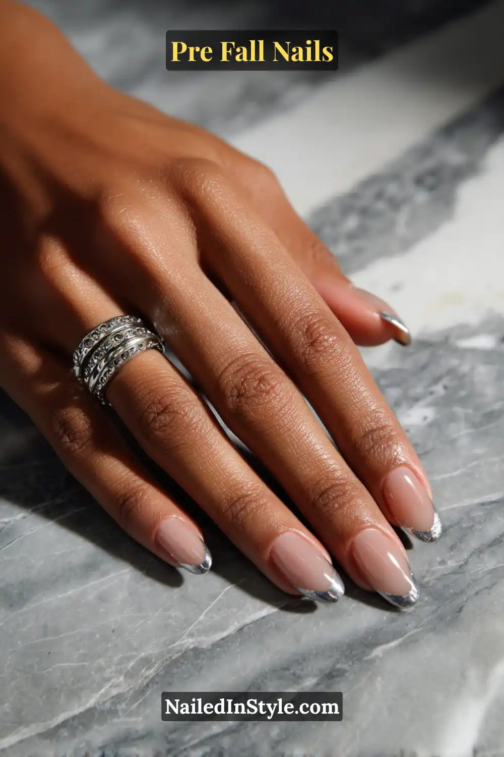 Almond nails with a sheer greige base framed by a tapered metallic slate outline, styled with silver rings on a marble background.