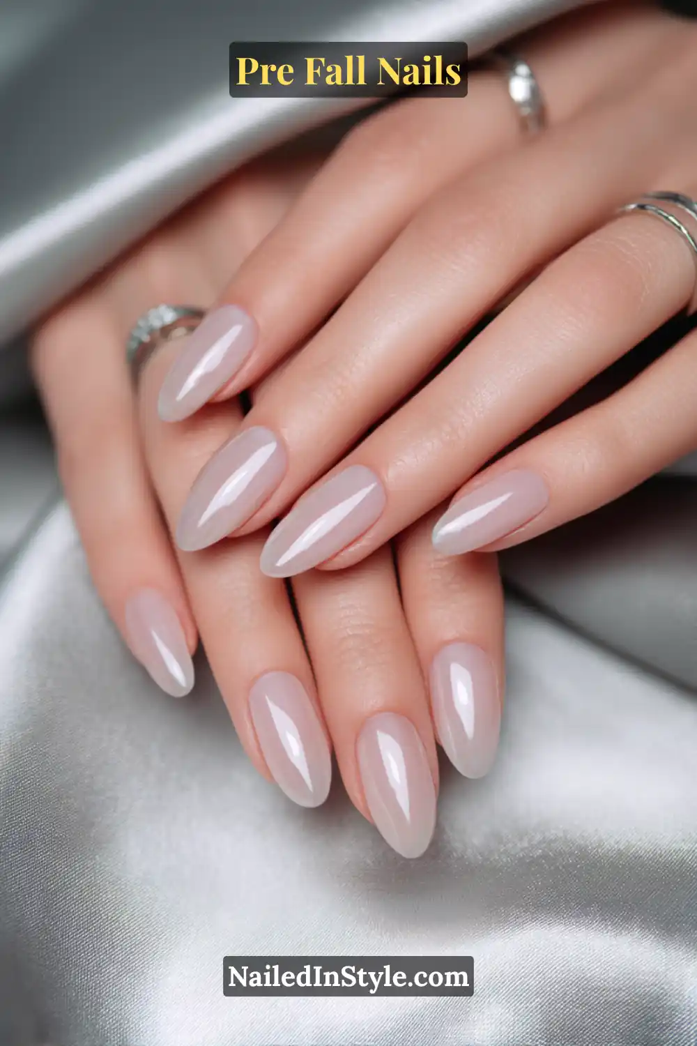 Oval nails with a translucent dove-grey sheer finish, allowing the natural nail warmth to show through, styled with silver rings on soft satin fabric.
