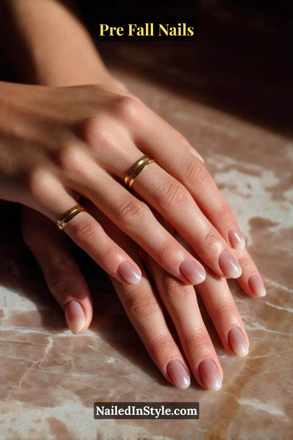 Short almond nails with a soft walnut-brown French blur, blending seamlessly into a sheer nude base, styled with gold rings on marble surface.