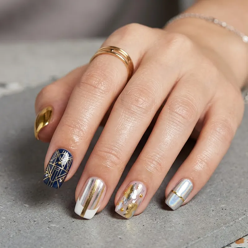 A professional close-up of a hand featuring contemporary metallic accent March nails. The manicure showcases a mix of navy blue with intricate gold geometric lines, vertical silver and gold stripes on a white base, and artistic gold and silver foil patterns on a neutral background. The look is accessorized with gold rings and styled against a modern grey stone and marble surface.