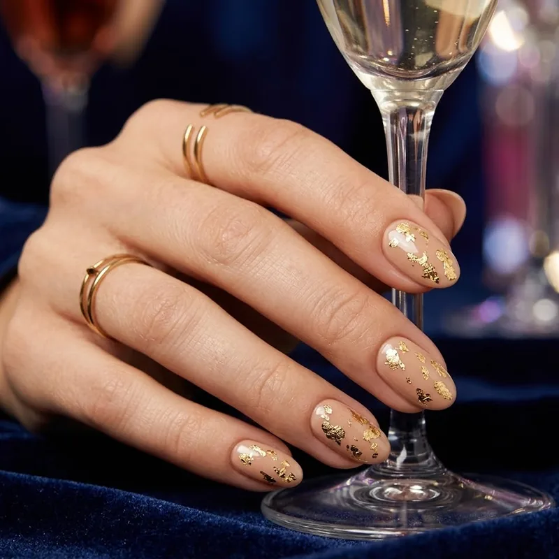 A hand holding a champagne glass, showcasing almond-shaped nude nails decorated with scattered gold leaf flakes for February nail ideas.