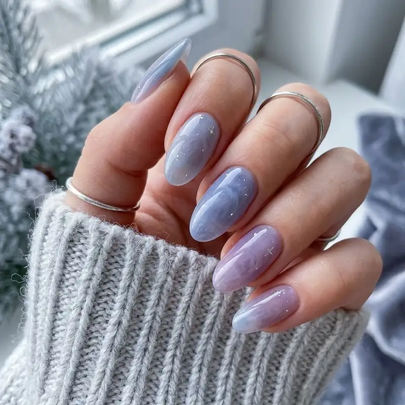 A hand displaying glossy almond-shaped nails with a sheer, marbled blend of soft blue and purple polish accented with tiny silver stars for February nail ideas.
