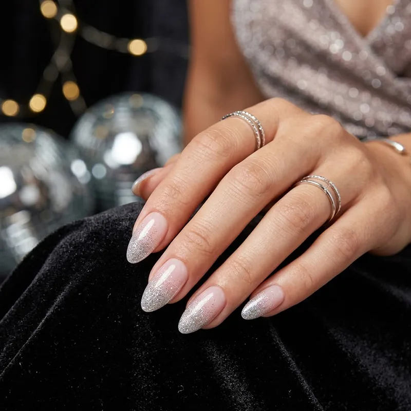 Prom Nails: Your Ultimate Guide to Stunning Styles and Techniques 2026 14 A professional close-up of almond-shaped prom nails featuring a subtle silver glitter gradient on a soft nude base, accessorized with silver rings against a glamorous backdrop of disco balls and sequins.