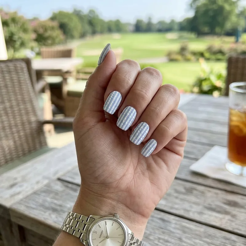 A high-resolution close-up photo of squoval nails painted with a light blue and white seersucker striped texture, wearing a luxury silver watch at a country club veranda.
