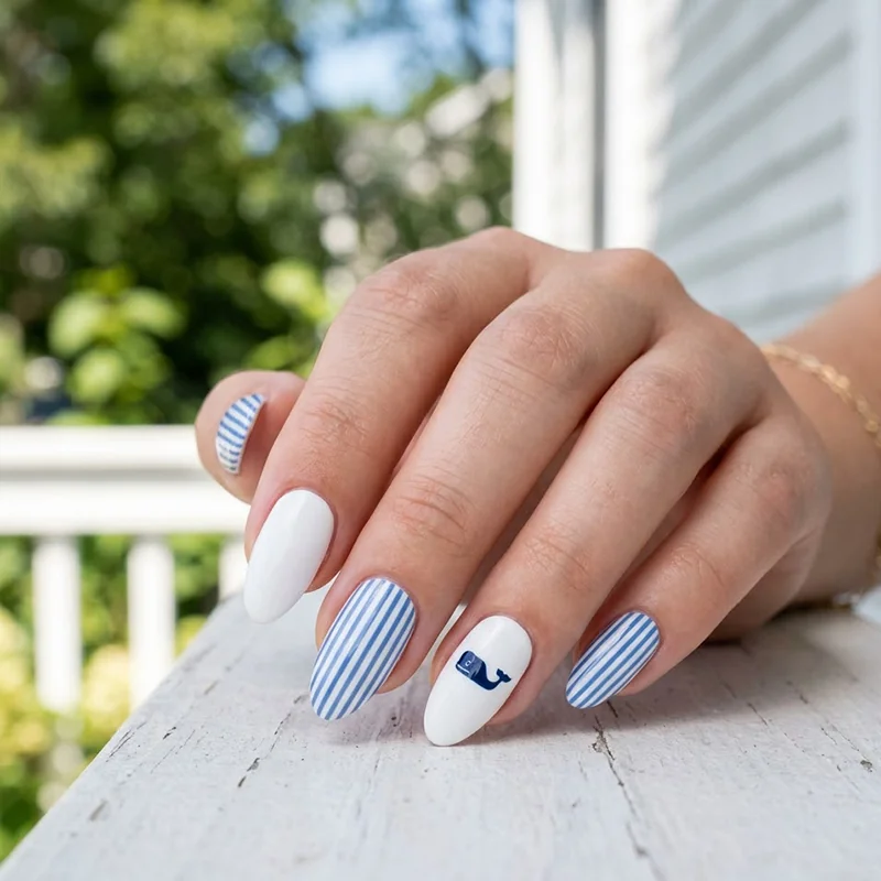 A professional close-up photo of almond-shaped preppy summer nails. The manicure features solid white nails, light blue and white vertical stripes, and a white accent nail on the ring finger with a navy blue whale logo decal. The hand rests on a white wooden porch railing with a blurred outdoor summer background.