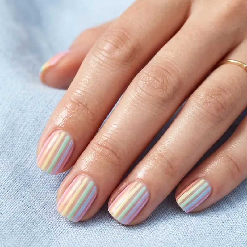 Close-up photograph of short, neat fingernails painted with precise vertical pastel rainbow stripes (mint, blue, yellow, pink) over a light base, holding a piece of light blue denim fabric. A delicate gold ring is visible on the index finger.