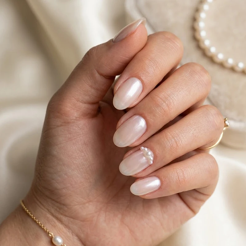 High-quality beauty photograph of medium oval nails with a sheer iridescent pearl base and delicate pearl bead accents near the cuticle of the ring finger.
